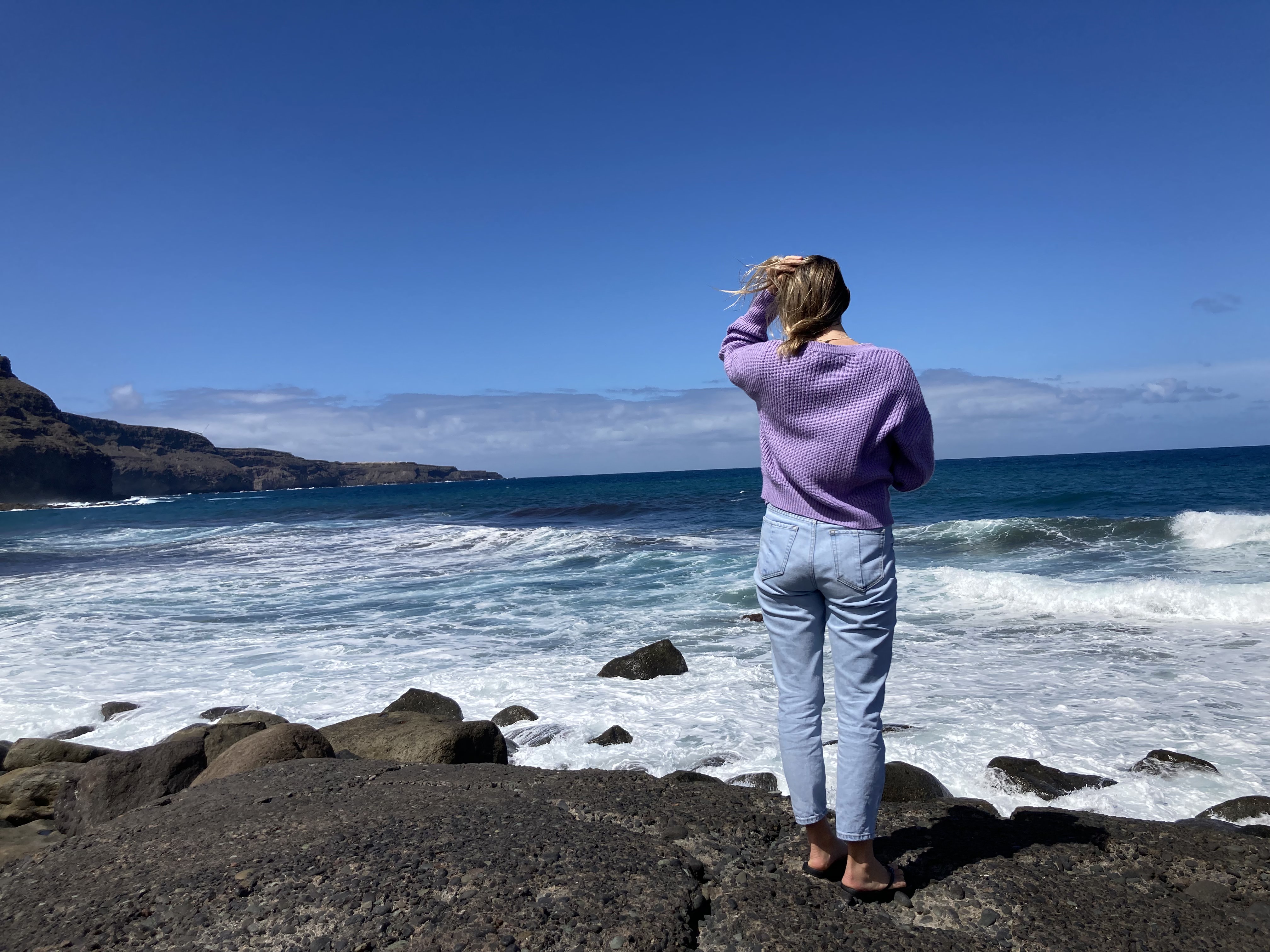 A person standing on rocky shore looking out at the ocean under a clear blue sky.
