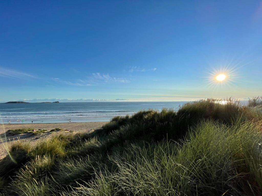 A scenic view of a beach framed by grass and under a clear blue sky, as the sun sets on the horizon.