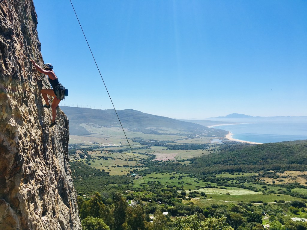 A person climbing a rocky cliff with a scenic view of rolling hills and coastline in the background.